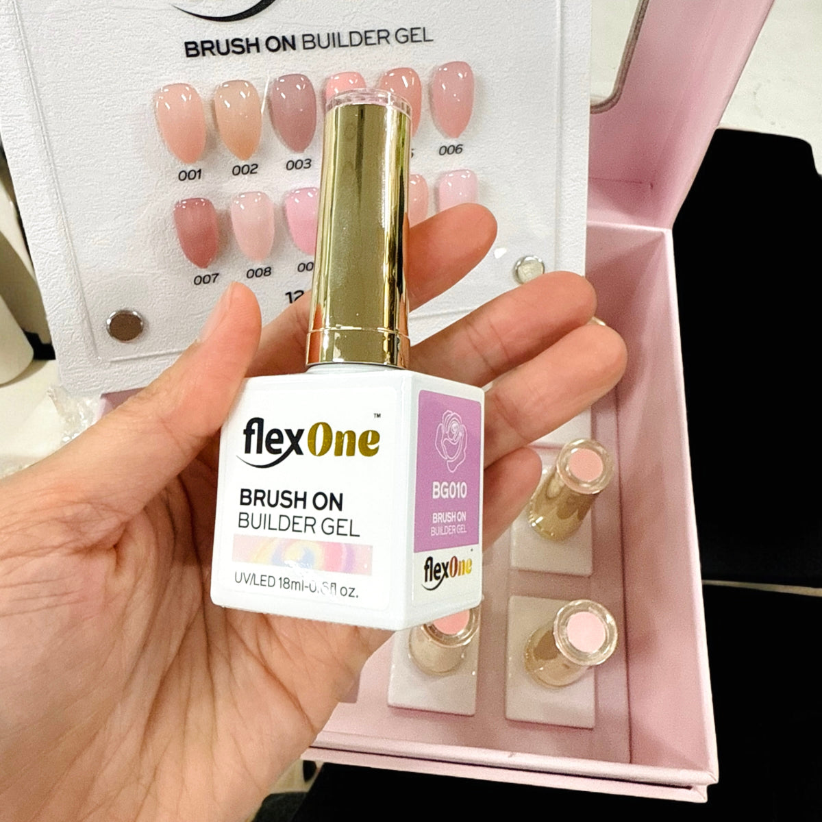 Hand holding a bottle of flexOne Brush On Builder Gel with a display of nail samples in the background.